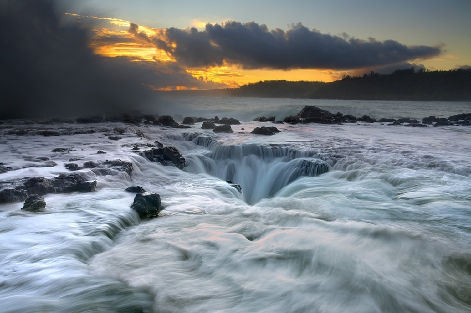 A dramatic seascape in Hawaii features turbulent waters flowing over rocks, illuminated by a vibrant sunset and dramatic clouds, showcasing the beauty of nature.