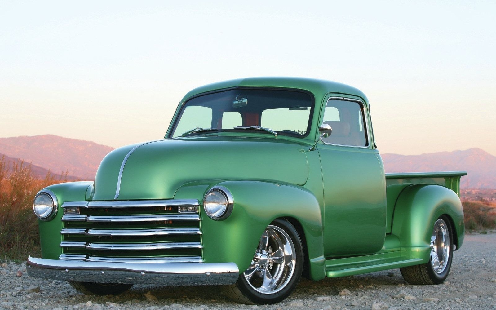 A green Chevrolet pickup truck parked on a dirt path with mountains in the background during sunset.