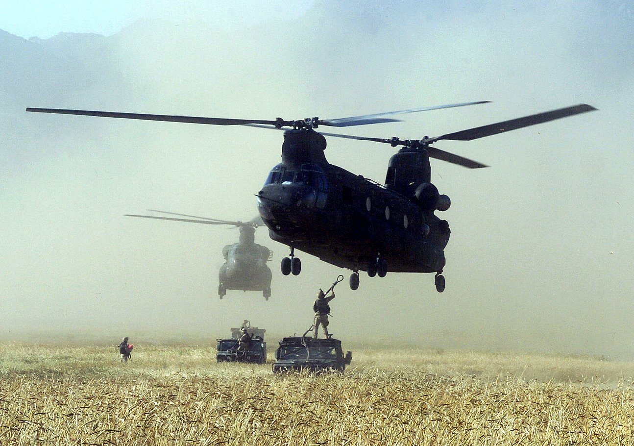 A soldier rappels from a Boeing CH-47 Chinook helicopter during a military operation in a dry, open field with another helicopter and vehicles nearby.