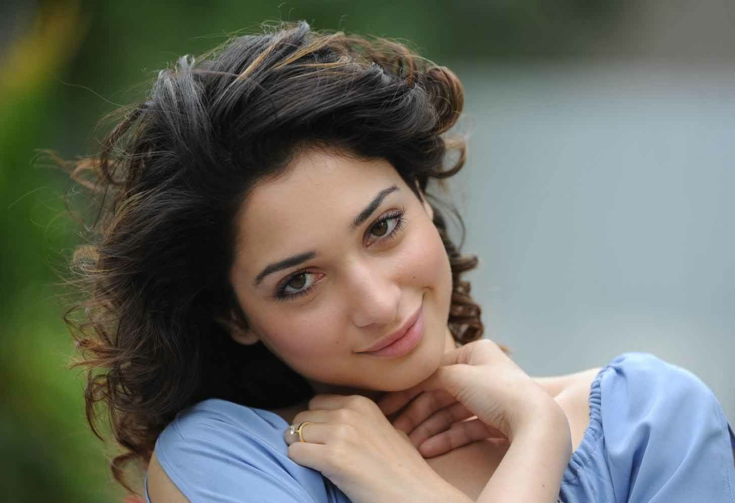Close-up portrait of celebrity Tamannaah Bhatia with curly hair, wearing a light blue top and posing with hands near her neck against a blurred background.