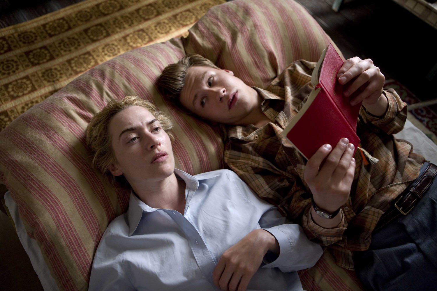 Scene from the movie The Reader featuring David Kross and Kate Winslet lying on a bed, with Kross holding and reading a red book.