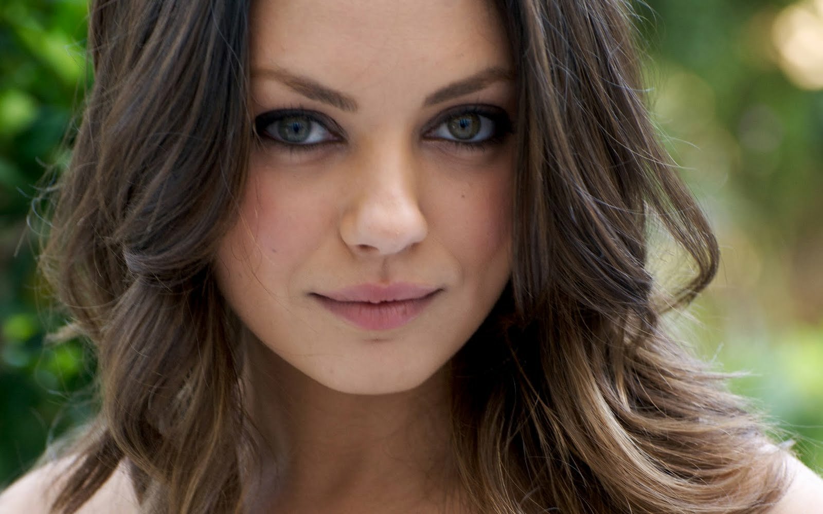 Close-up portrait of celebrity Mila Kunis with soft waves in her hair, striking blue eyes, and a natural makeup look against a blurred outdoor background.