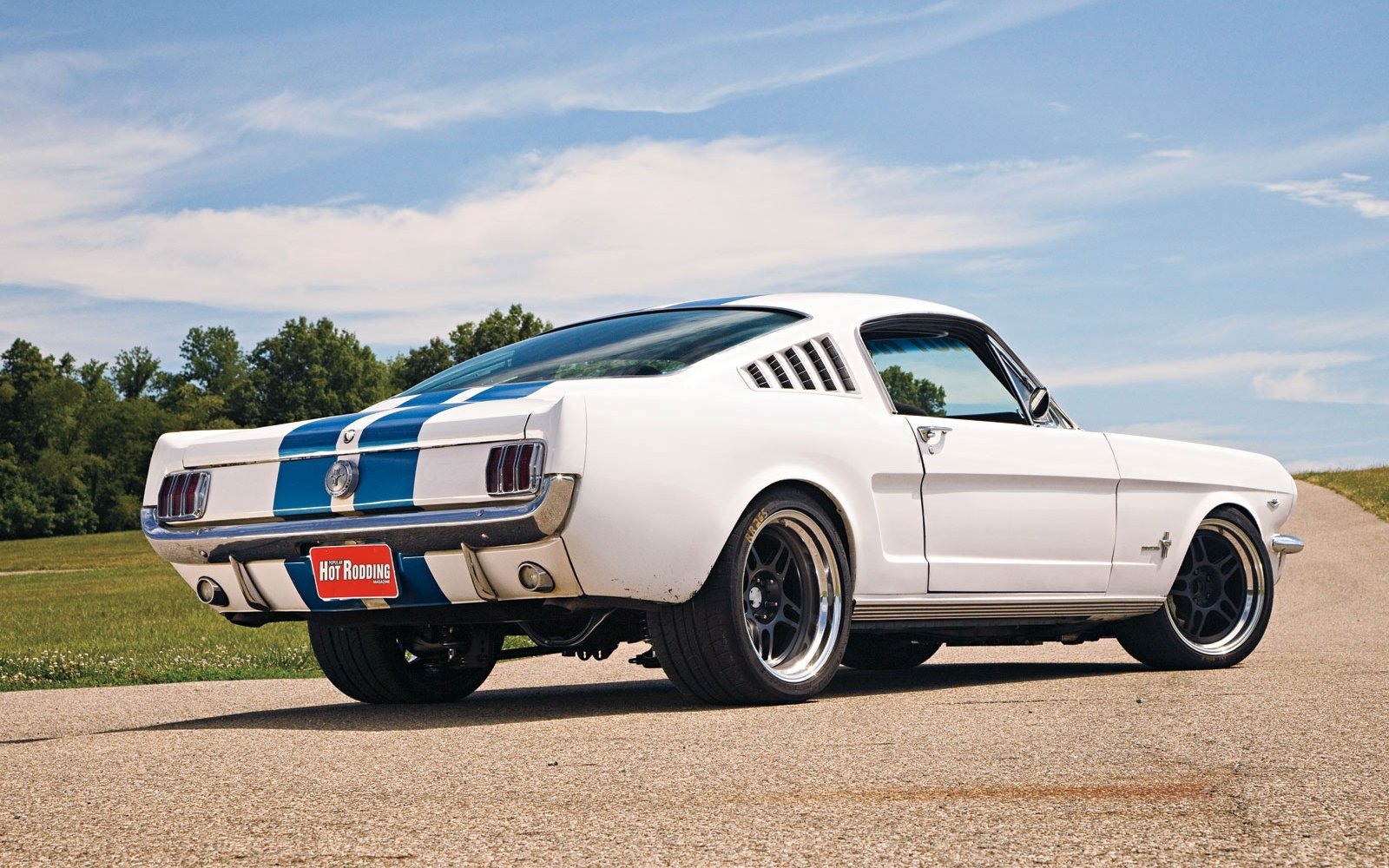 White Ford Mustang Fastback muscle car with blue racing stripes parked on a paved surface under a clear sky.