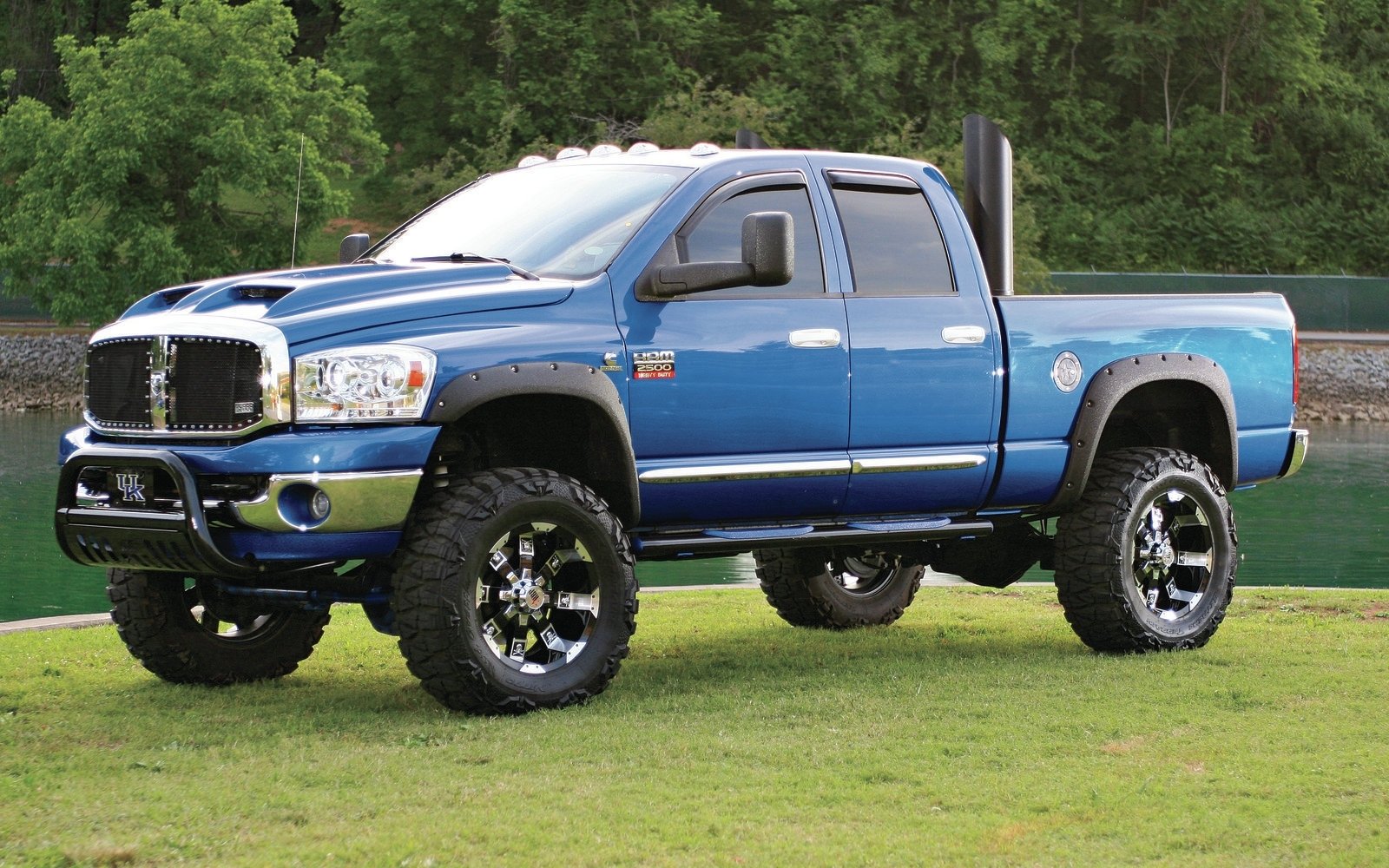 A blue Dodge Ram 2500 truck with oversized tires is parked on green grass, surrounded by trees and water, showcasing its robust design and powerful presence.
