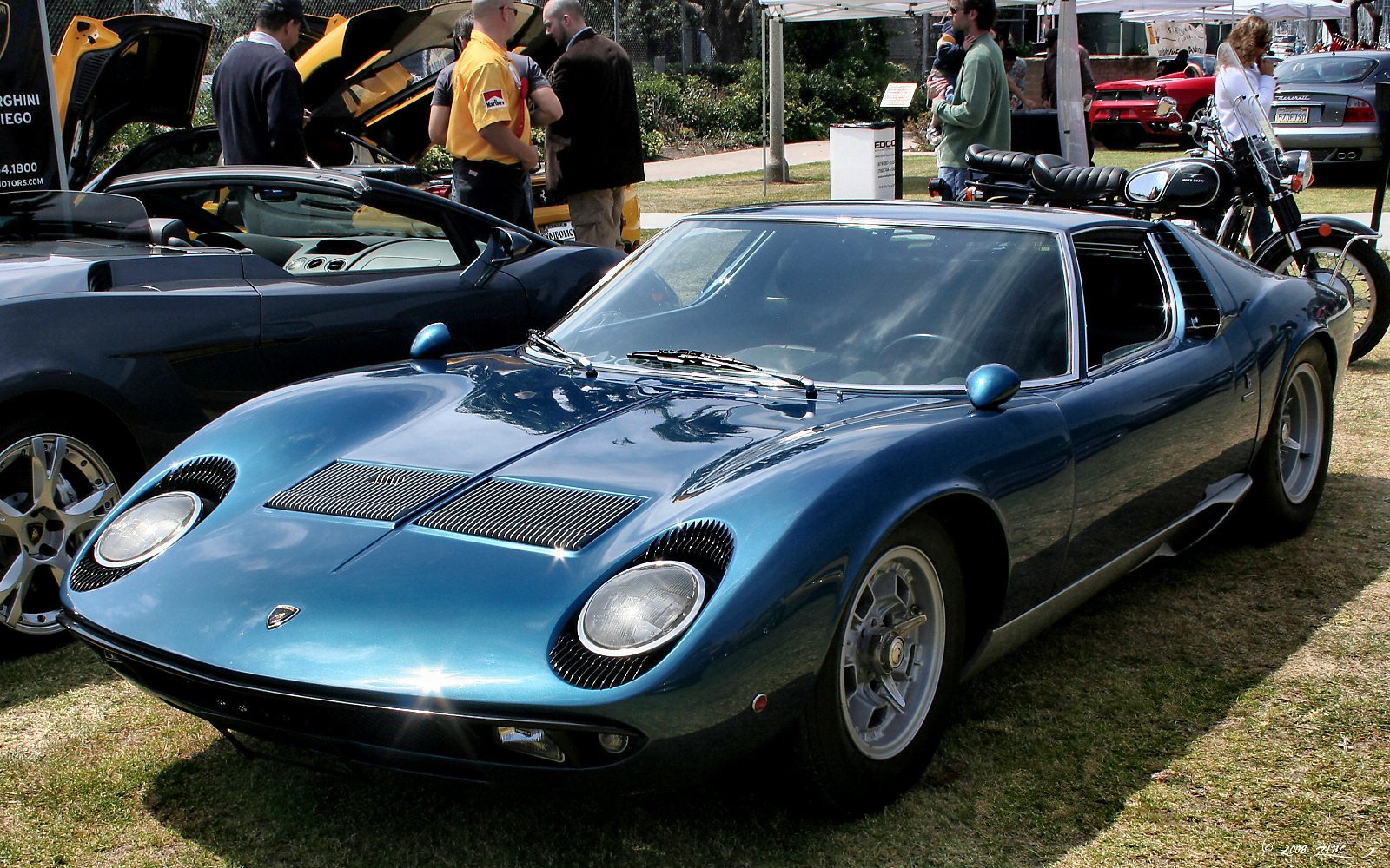 A sleek blue Lamborghini Miura sports car is displayed outdoors at a car show, surrounded by spectators and other vehicles.