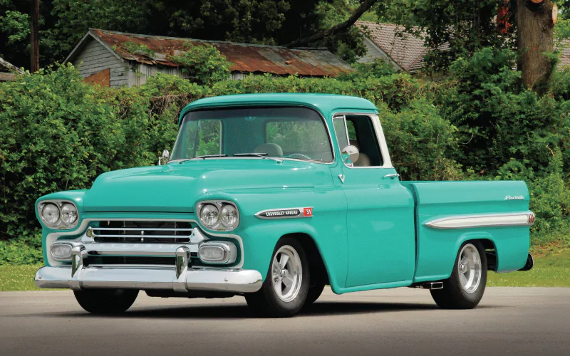 A turquoise 1959 Chevrolet Apache pickup truck parked on a paved surface with greenery and rustic buildings in the background.