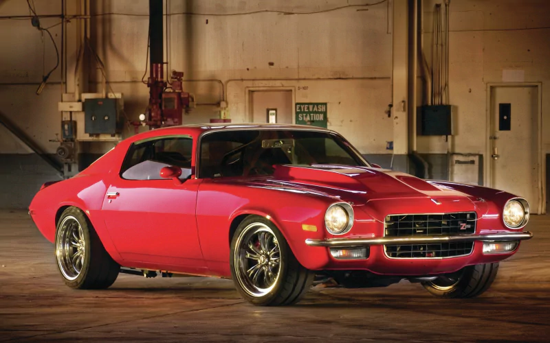 A red Chevrolet Camaro Z28 muscle car parked inside a spacious industrial garage with warm lighting highlighting its sleek design.