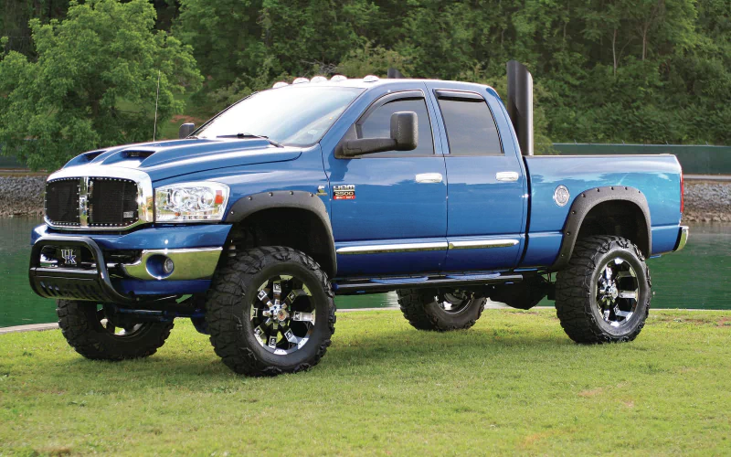 A blue Dodge Ram 2500 truck with oversized tires is parked on green grass, surrounded by trees and water, showcasing its robust design and powerful presence.