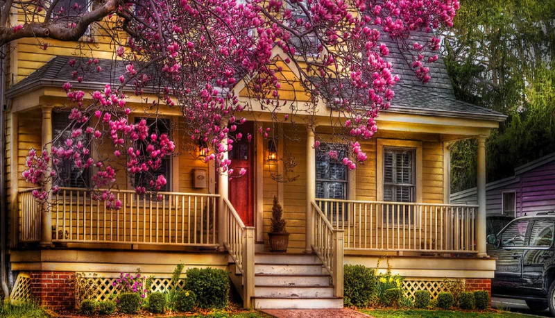 A charming yellow house with a front porch is surrounded by vibrant pink blossoms, creating a picturesque scene of man-made beauty in a serene neighborhood.
