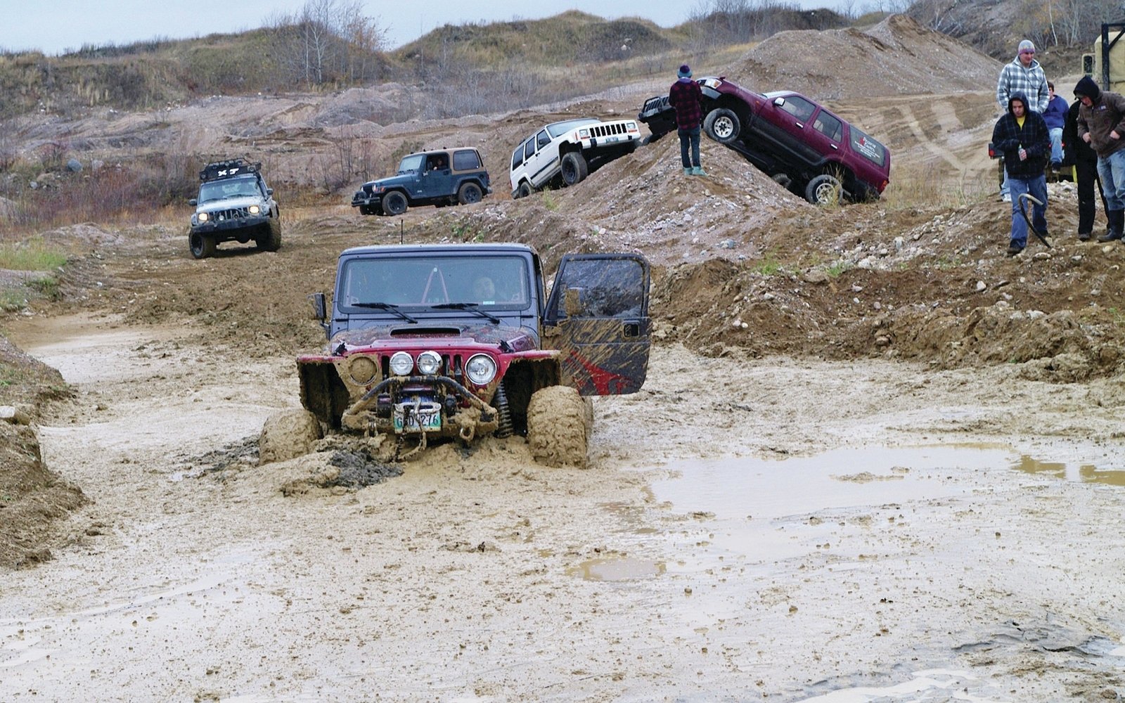 A group of Jeep vehicles navigate muddy, uneven terrain while people observe and assist during off-road driving activities.