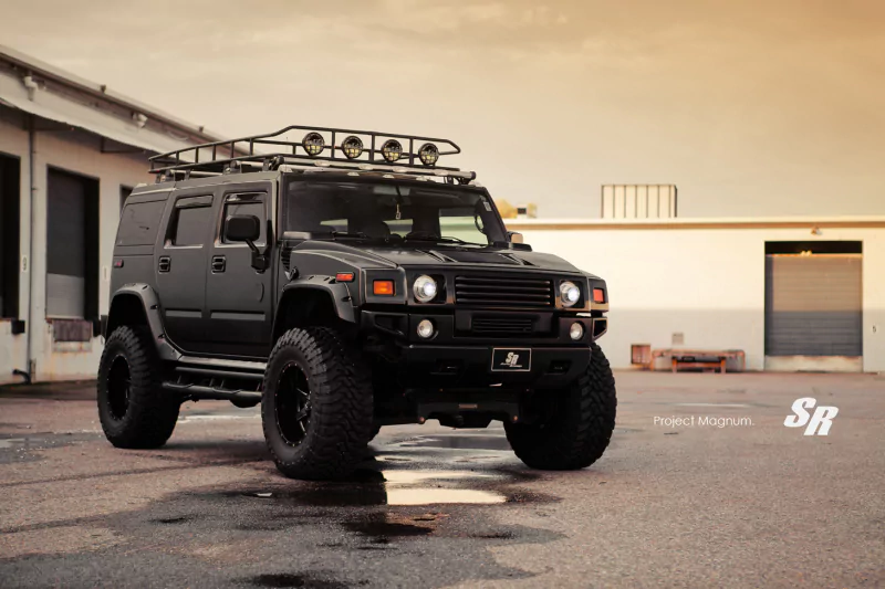 A black Hummer H2 vehicle with off-road tires and roof lights is parked on a wet concrete surface near industrial buildings during sunset.