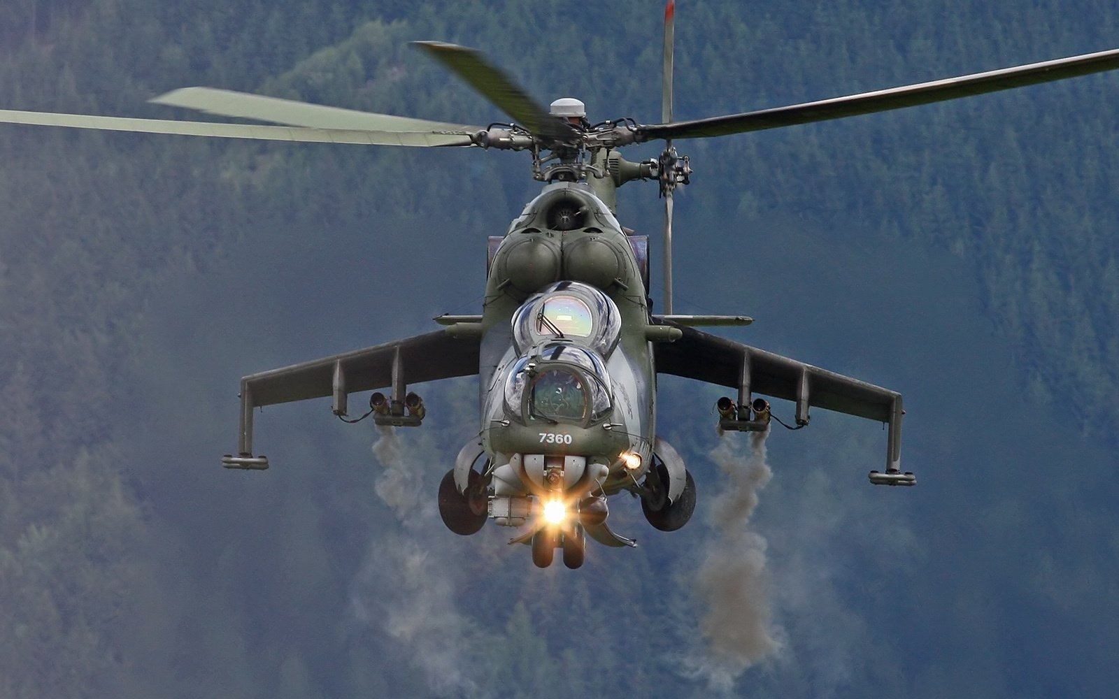 A military helicopter flies forward with its rotors spinning and smoke trailing from its weapons, set against a backdrop of mountains and clear skies.