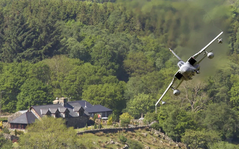 A military Panavia Tornado jet flies low over a forested area near a house nestled among trees.