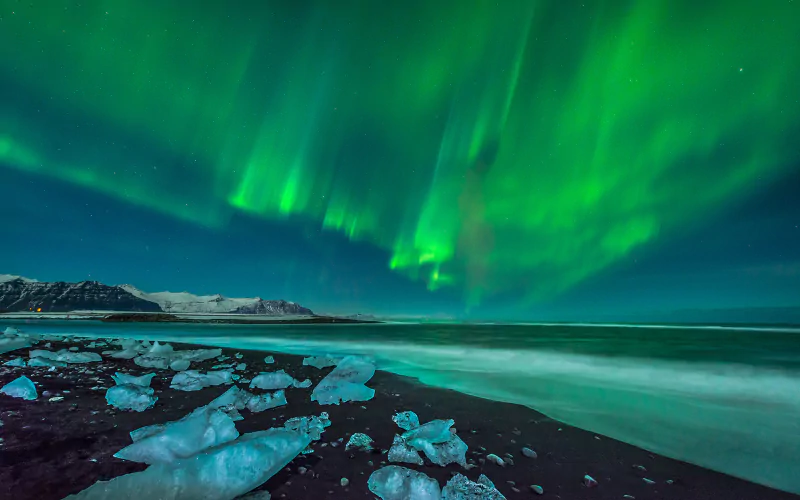 A stunning display of the aurora borealis lights up the night sky with vibrant green hues, casting a magical glow over a serene beach and scattered ice formations.