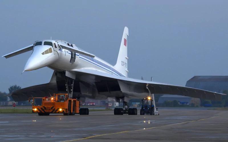 Tupolev Tu-144 supersonic airliner vehicle on an airport ramp at dusk, flanked by ground service vehicles and tow tractors.