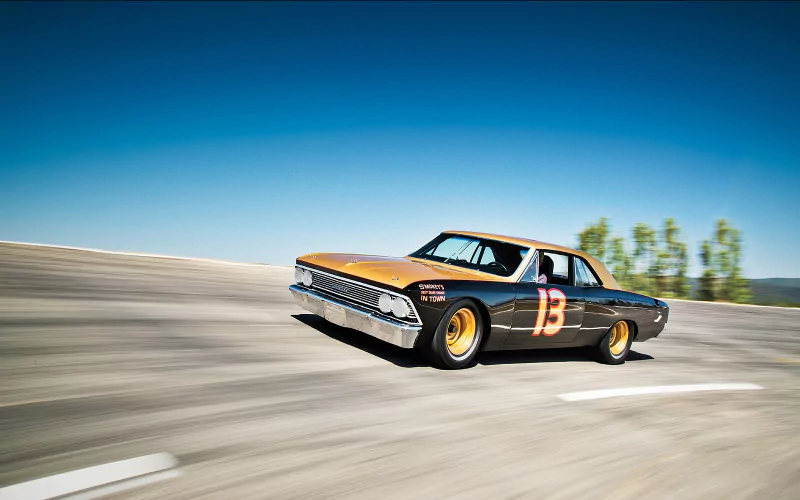 A 1966 Chevrolet Chevelle with the number 13 races on a curved track under a clear blue sky.