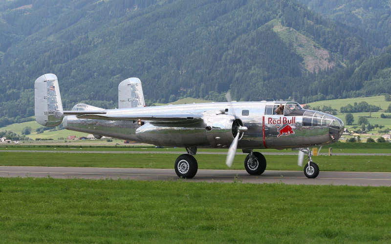 A vintage aircraft vehicle with Red Bull branding taxis on a runway surrounded by green fields and forested hills.