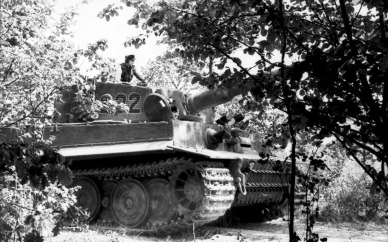 Black and white image of a Tiger I military tank partially concealed by trees and foliage in a forested area.