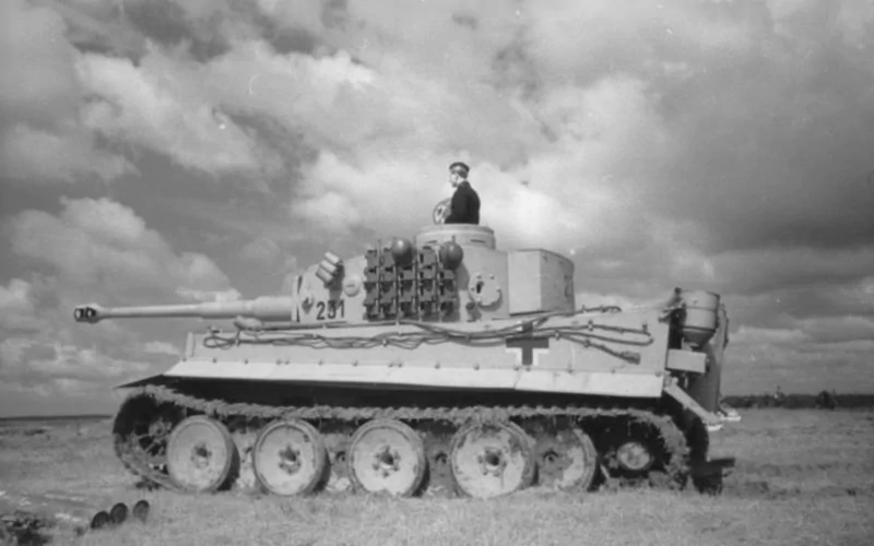 Black and white photo of a Tiger I military tank with a soldier visible in the open turret, positioned on open terrain under a cloudy sky.