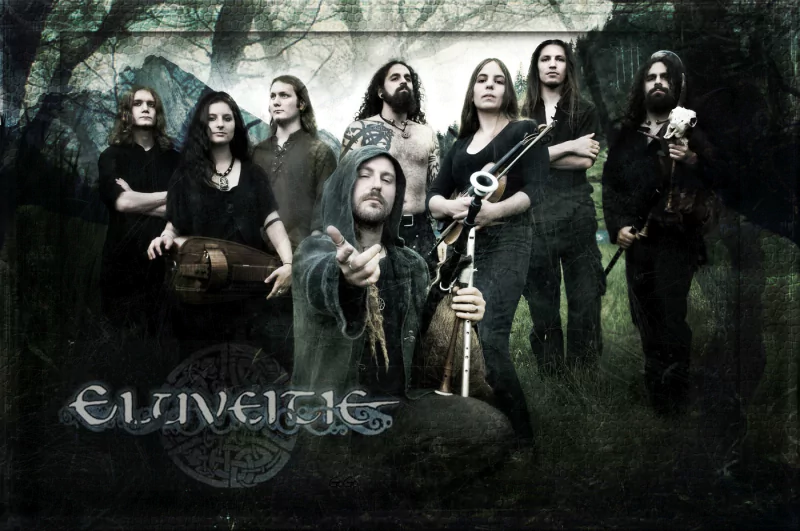 A group photo of the folk metal band Eluveitie, with members posing in a dark, forested background holding traditional musical instruments.