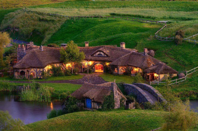 A picturesque Hobbiton scene featuring cozy, man-made houses with thatched roofs, surrounded by lush greenery and a tranquil pond in the foreground.