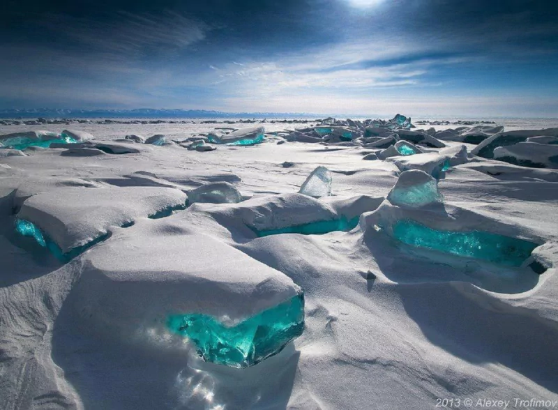 A serene landscape in Russia showcases turquoise ice formations scattered across a snowy expanse, illuminated by a bright sky, capturing the beauty of winter nature.