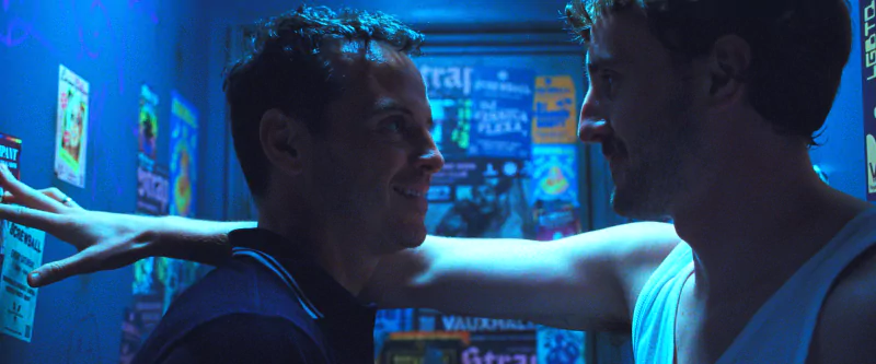 A scene from the movie All of Us Are Strangers featuring two male characters sharing a close moment in a dimly lit room with posters on the wall.