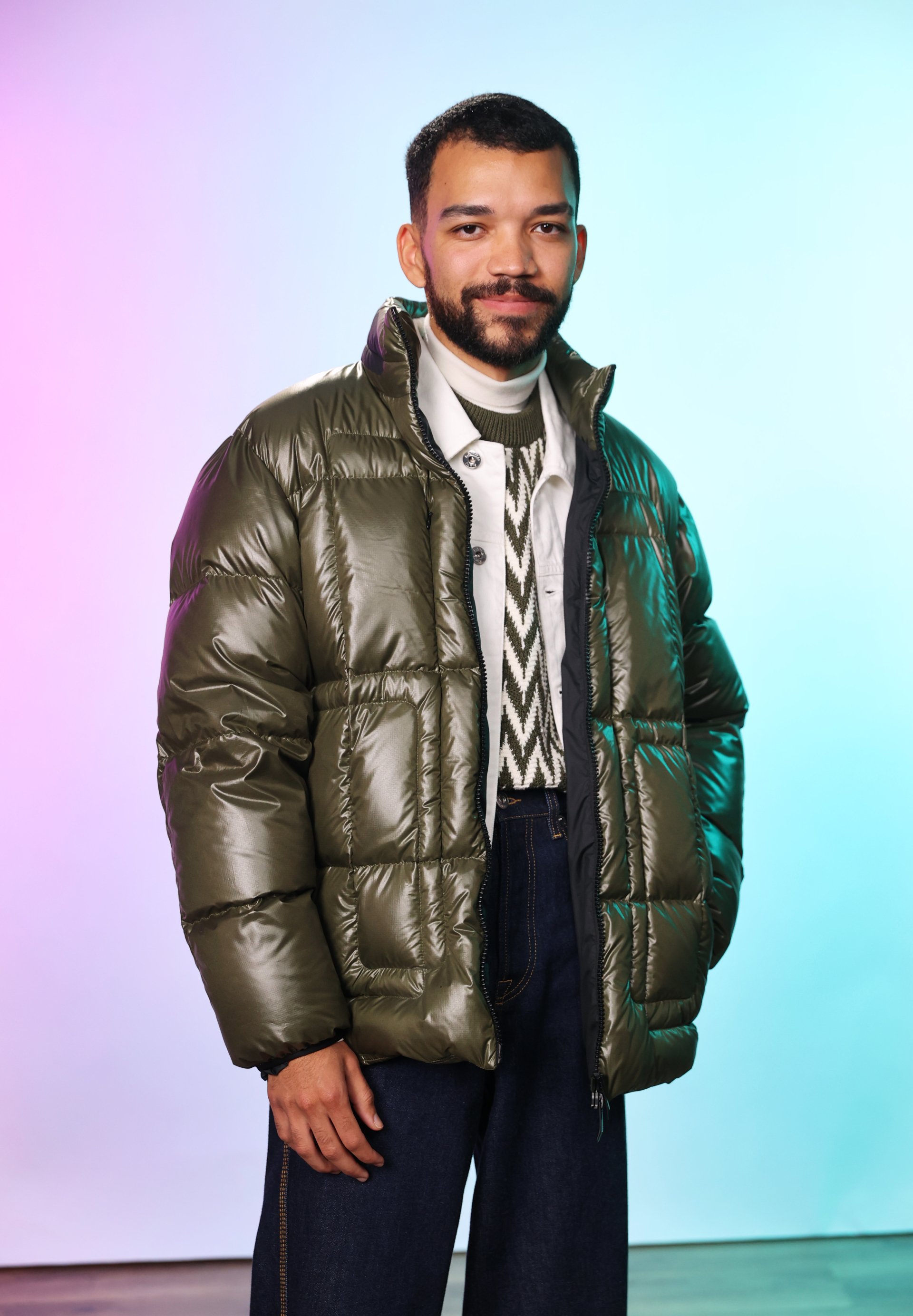 Man from 'The American Society of Magical Negroes' TV show wearing a puffy jacket and patterned sweater standing against a colorful background.