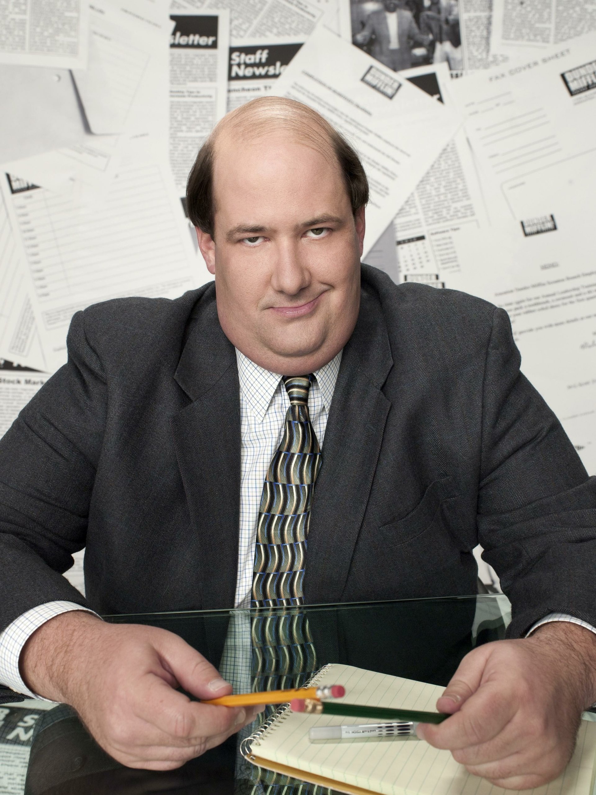 Actor from The Office (US) TV show sitting at a desk surrounded by papers, portraying a character at work.