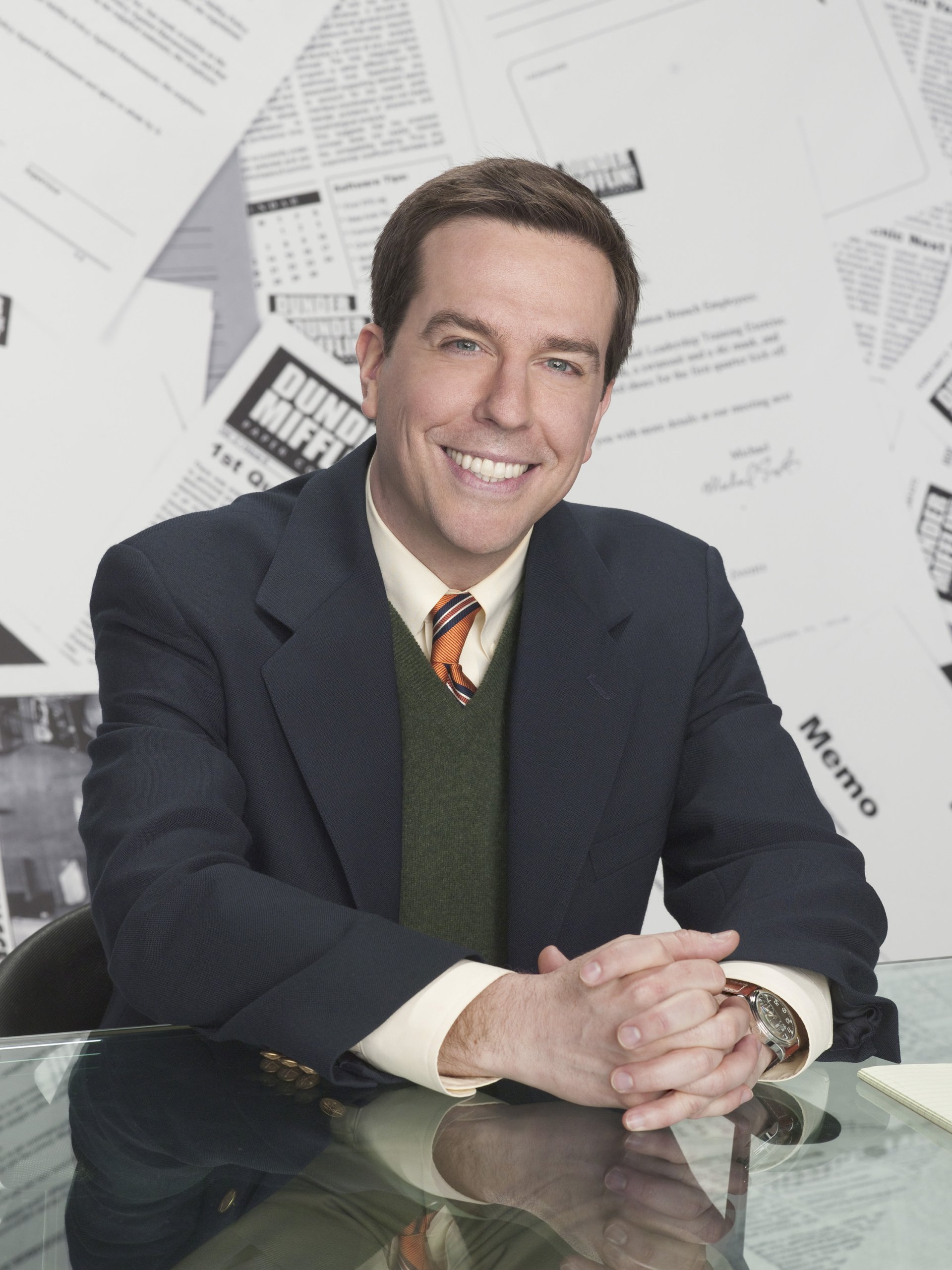 Smiling man in a suit with a striped tie posing at an office desk, surrounded by scattered papers, representing a scene from the TV show The Office.