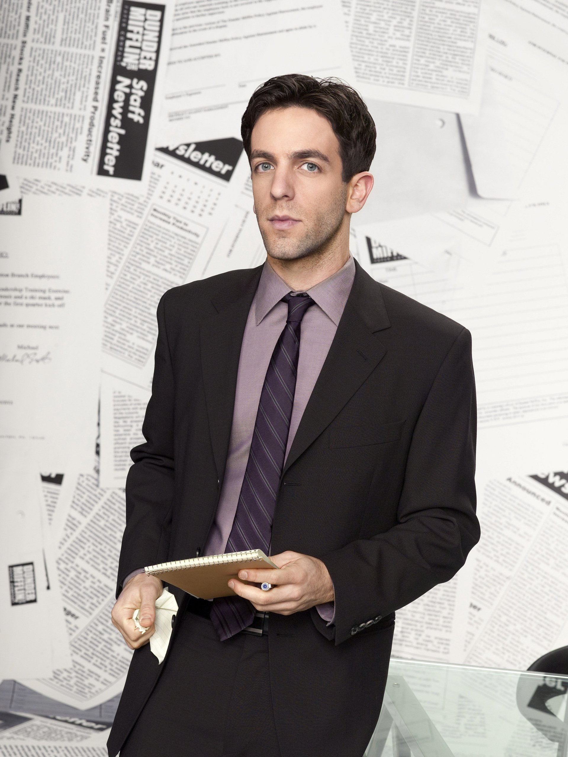 Professional-looking character from The Office (US version) standing with a clipboard in hand, against a backdrop of paper-covered walls, dressed in a sharp suit and tie.