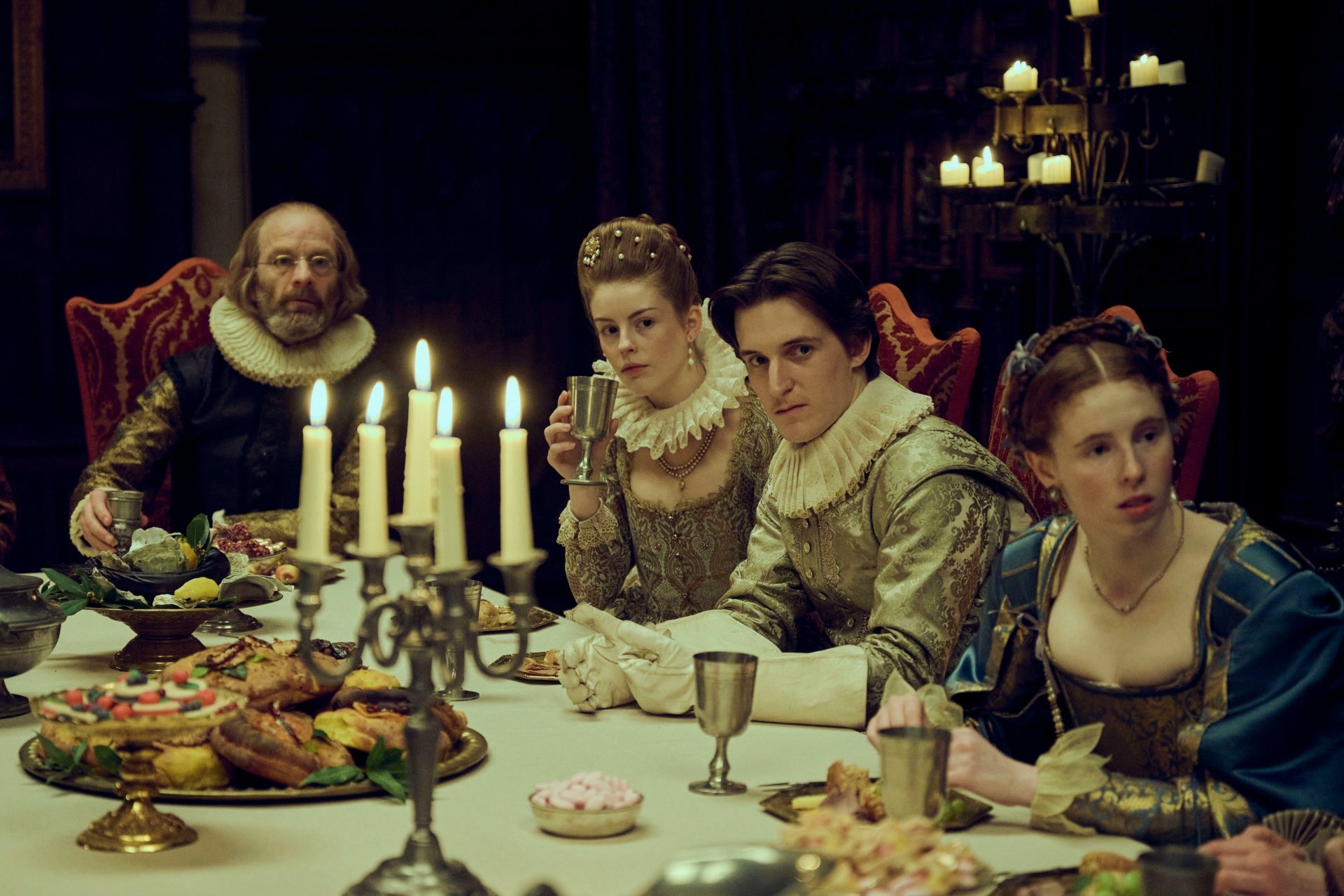 A scene from the TV show Mary & George featuring four characters dressed in historical costumes sitting at a banquet table lit by candles.