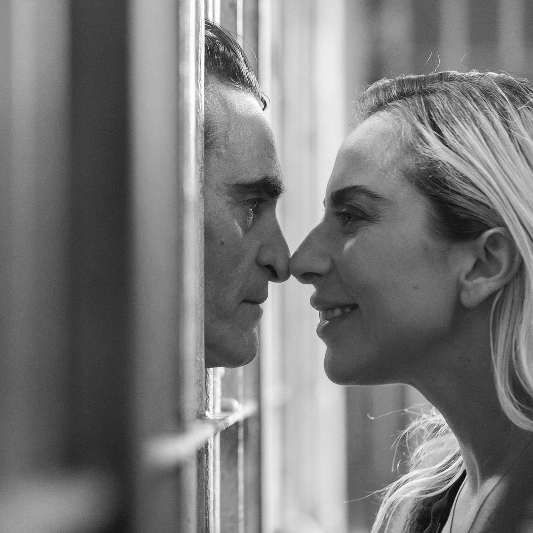 Black and white image of a man and woman facing each other with noses almost touching, separated by blurred vertical bars, suggestive of a scene from Joker: Folie à Deux.