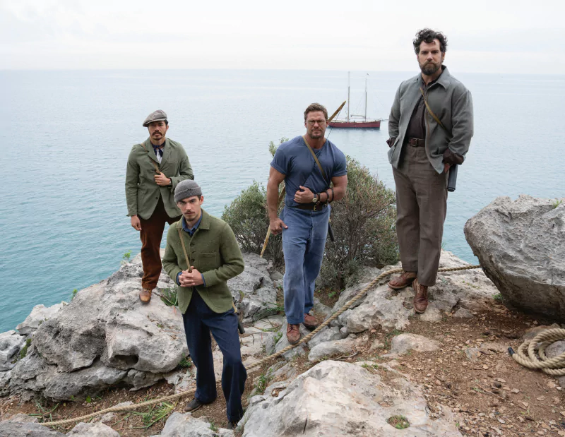 Four actors portraying soldiers from The Ministry of Ungentlemanly Warfare standing on a rocky coastline with the sea and a sailboat in the background.