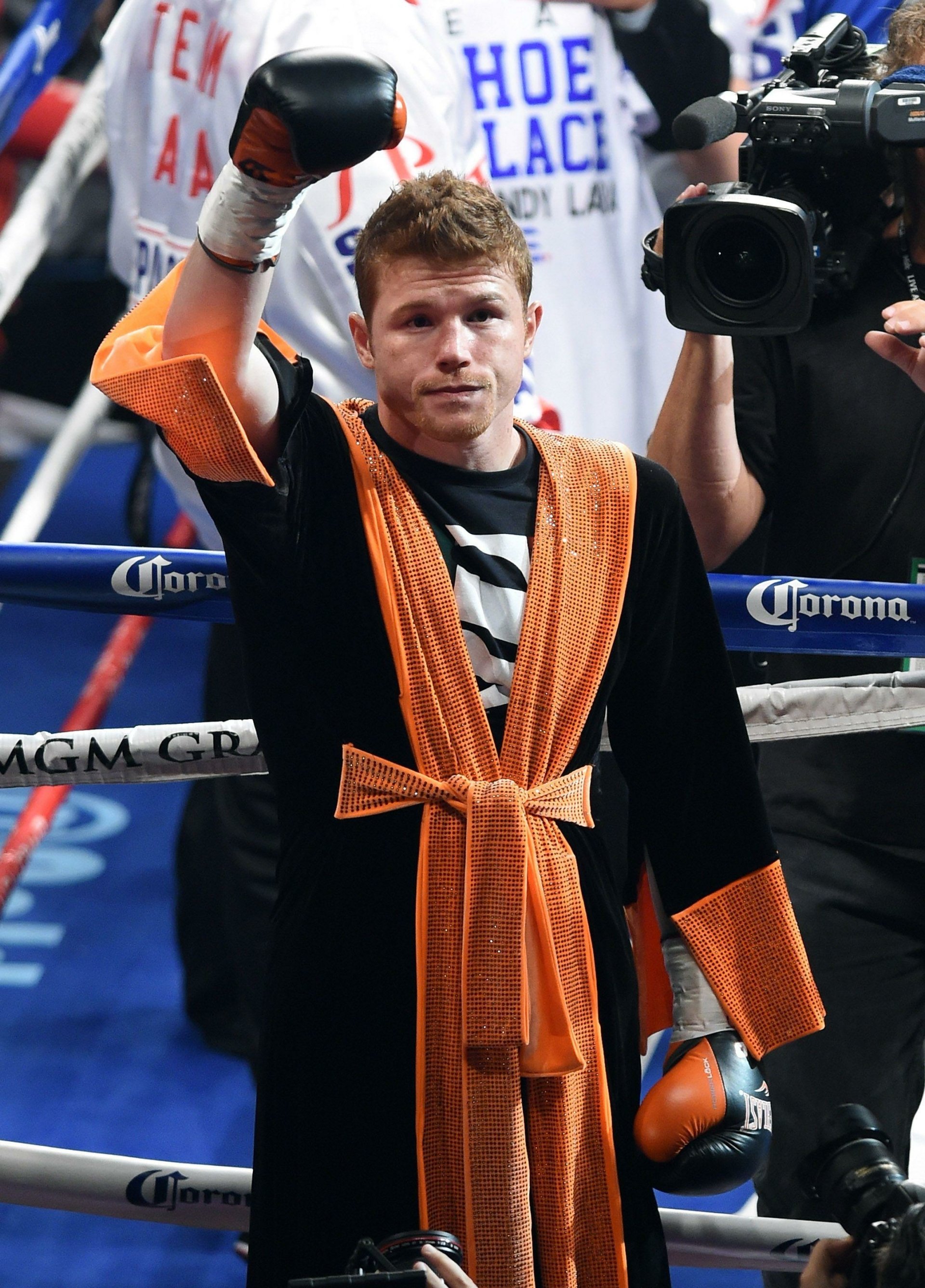 Canelo Álvarez in a boxing ring, sporting orange and black robe with raised fist, celebrating a win at an MGM Grand event.