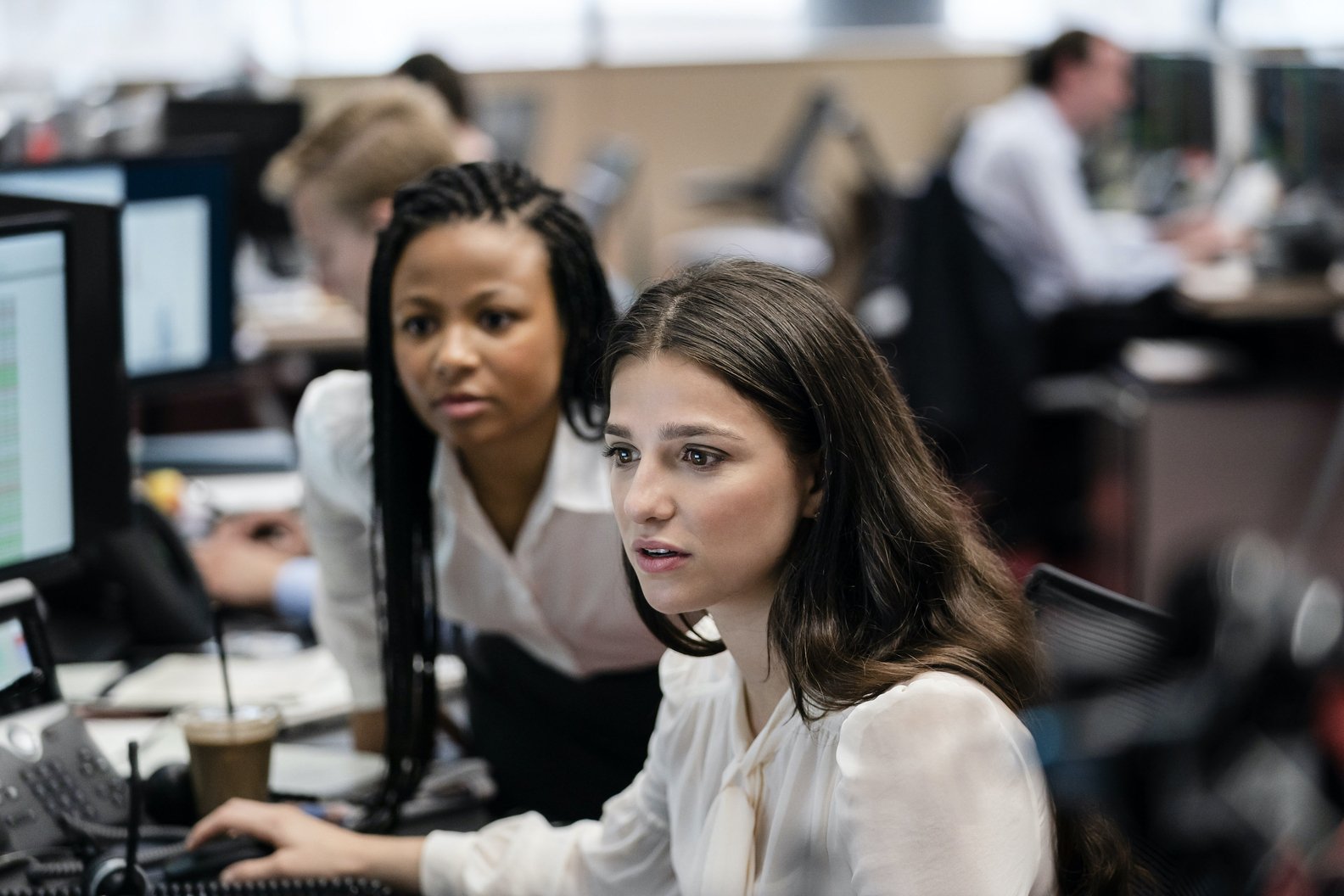 Two women focus intently on a computer screen in a bustling office environment, depicting a scene from the TV show Industry. Other employees work diligently in the background.