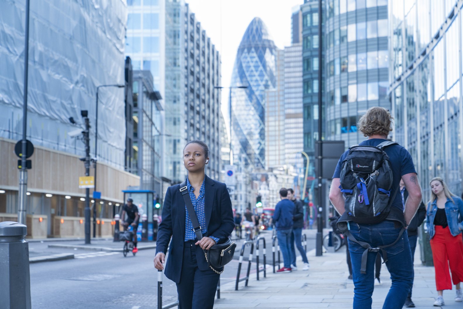 A street scene from the TV show Industry, featuring a woman in business attire walking among modern buildings, while pedestrians and cyclists share the busy urban environment.