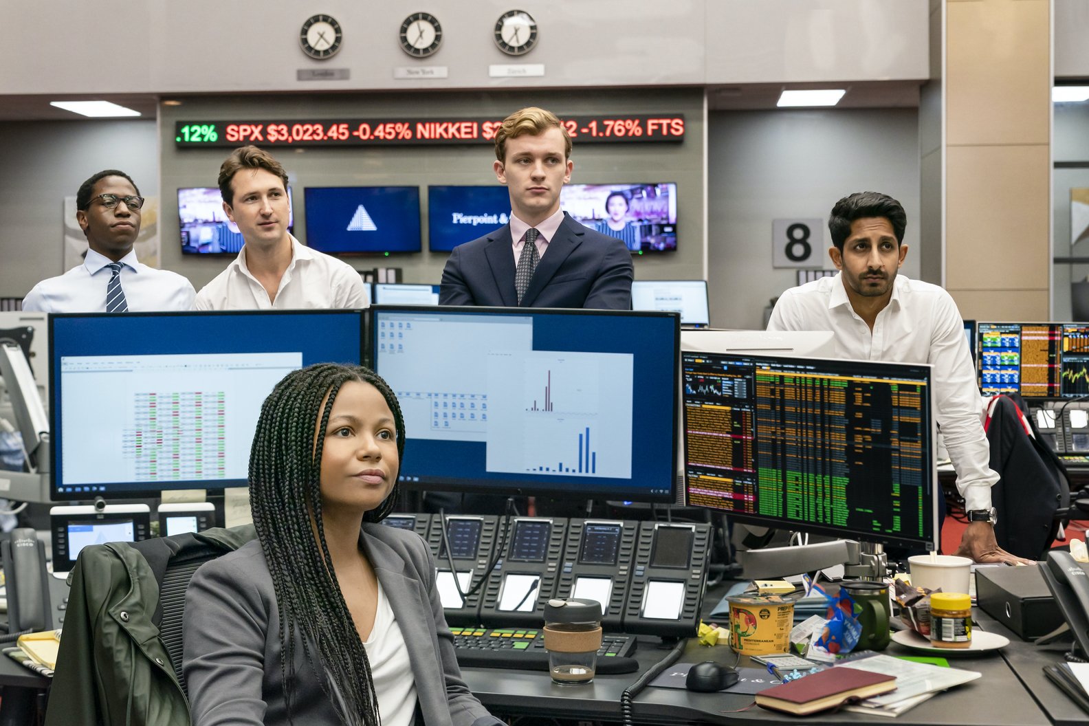 A scene from the TV show Industry, featuring a diverse group of young professionals in a busy trading room, focused on monitors displaying stock data and financial charts.