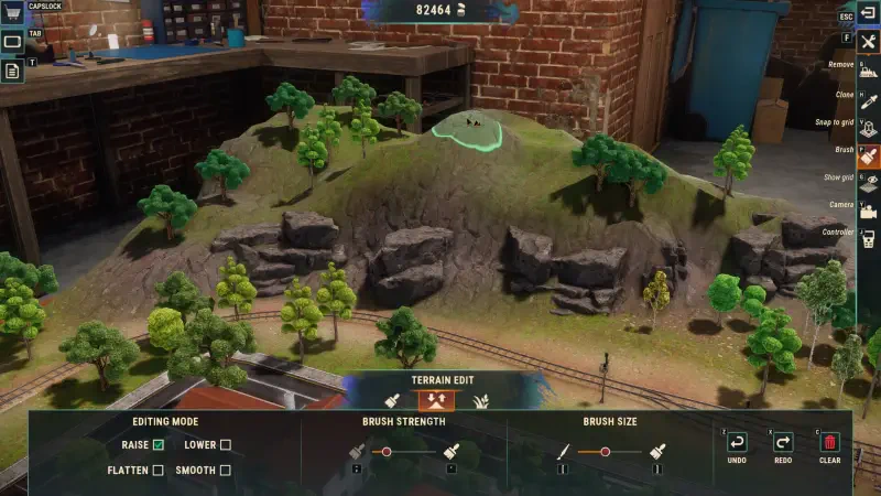 A detailed view of a Train Yard Builder video game scene, showcasing a miniature landscape with hills, rocks, and trees, set against a brick wall background.