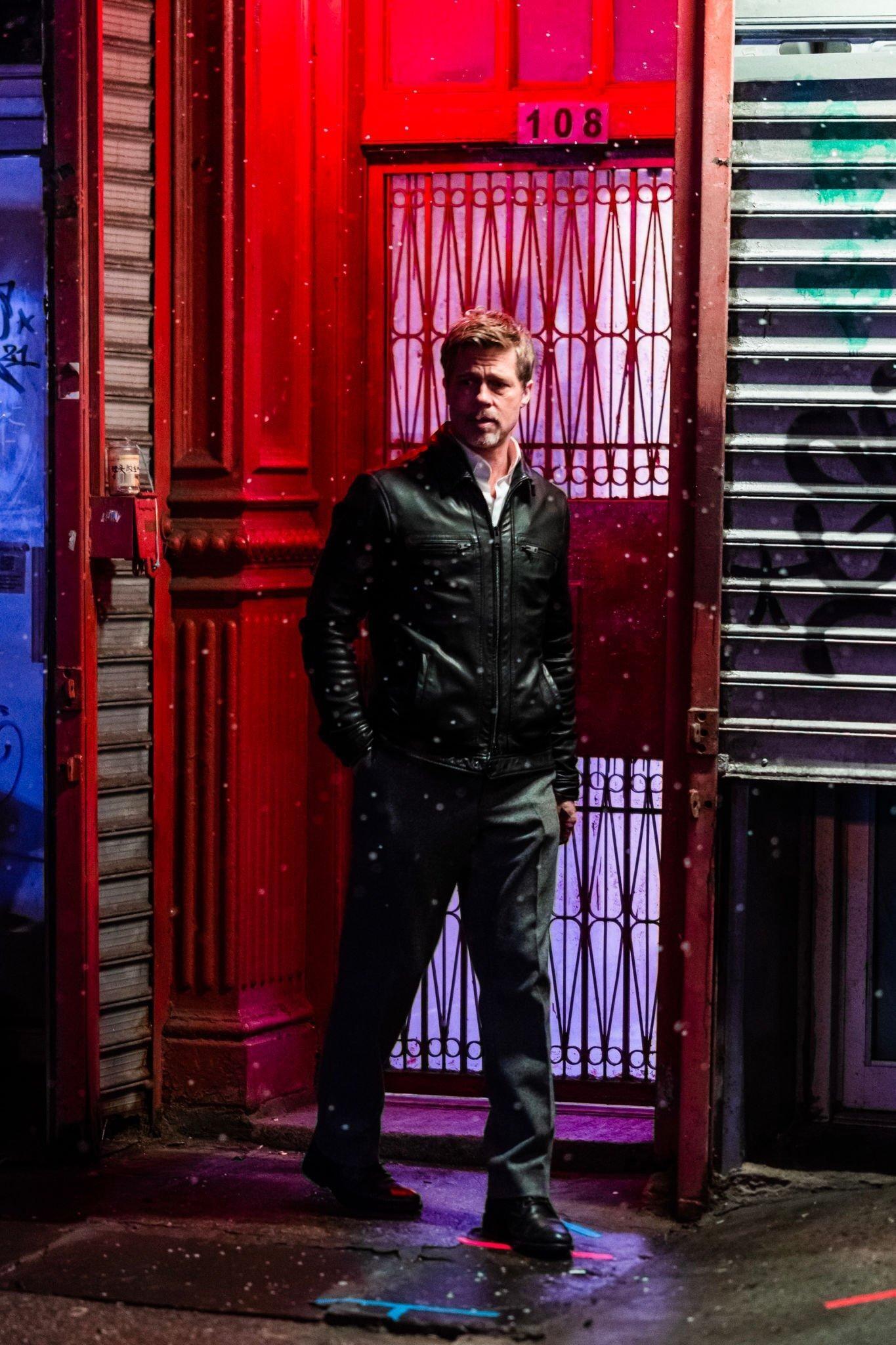A scene from the movie Wolfs (2024) featuring Brad Pitt standing in front of a vibrant red door, dressed in a black leather jacket against a nighttime urban backdrop.