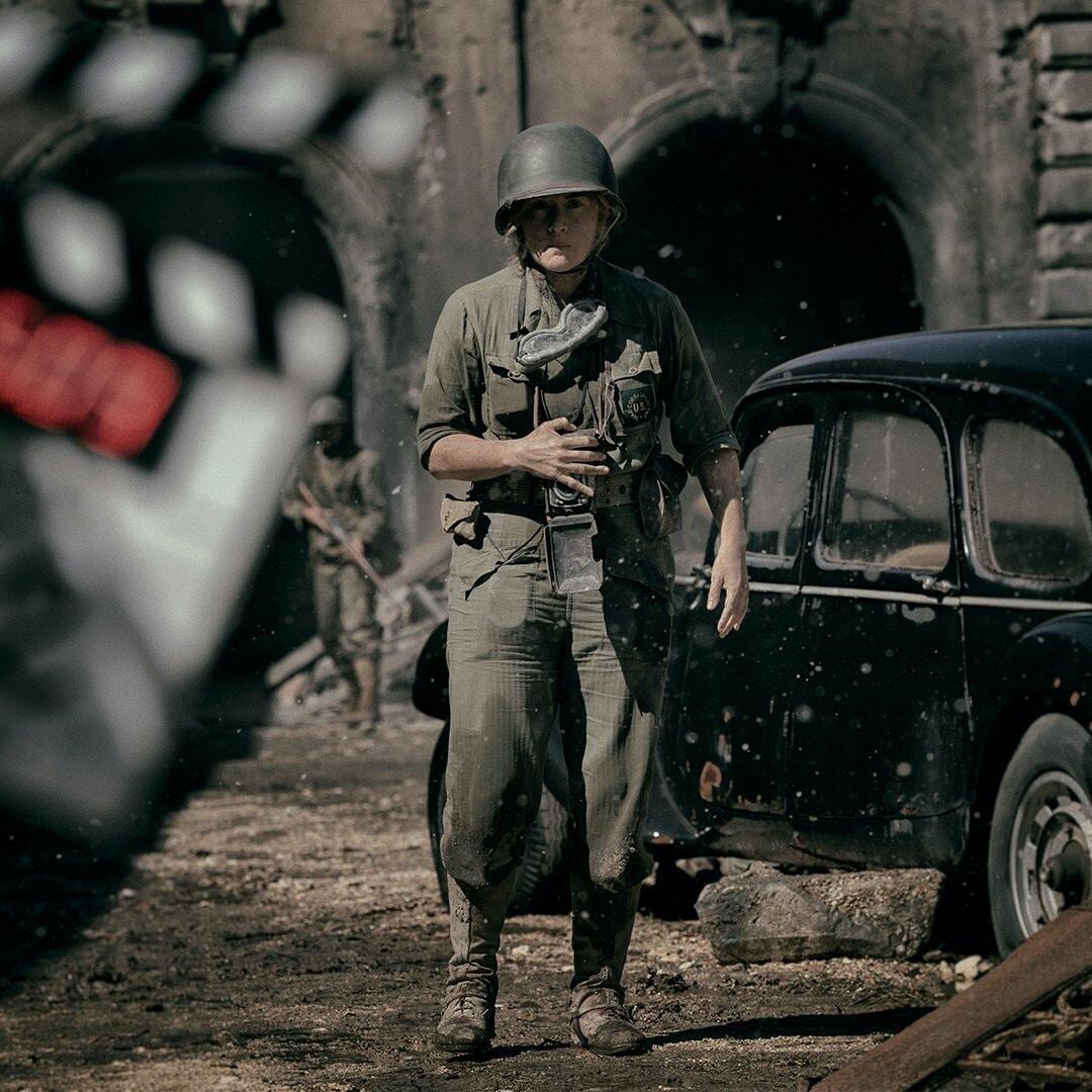 A soldier stands in a war-torn setting, with a clapperboard in the foreground, from the movie Lee (2023) starring Kate Winslet. The scene captures a dramatic moment of conflict.