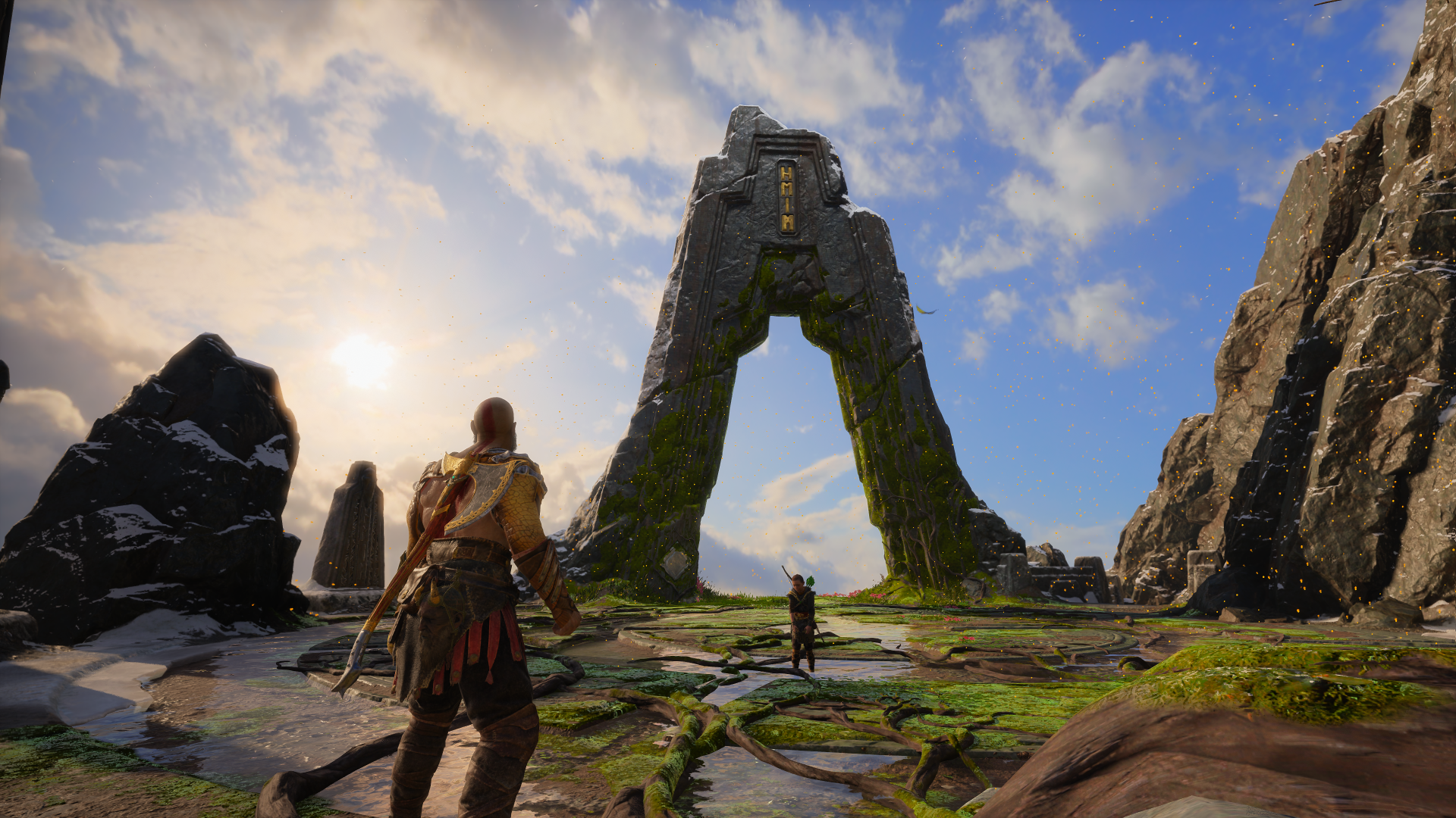 Kratos and Atreus stand before a towering moss-covered stone archway beneath a bright sky in a coastal, rocky Norse landscape — God of War scene.