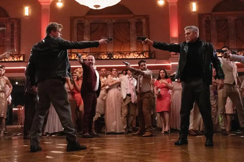 A tense scene from the movie Wolfs (2024), featuring two characters facing off with guns in a crowded ballroom, as onlookers watch in suspense.