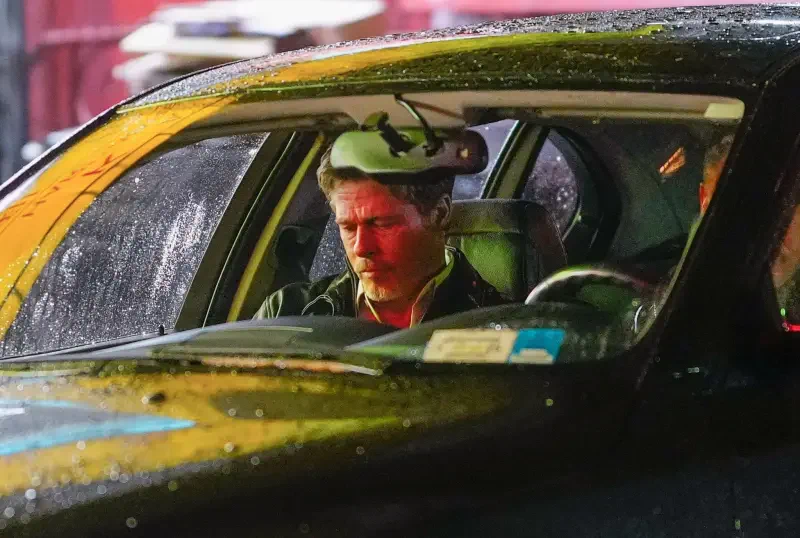 Brad Pitt is seated in a car, looking contemplative amid raindrops on the windshield, in a scene from the 2024 movie Wolfs.