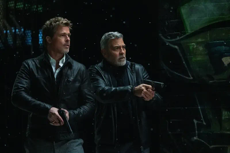 Brad Pitt and George Clooney action pose with guns in a scene from the movie Wolfs (2024), set against a dark, atmospheric backdrop.