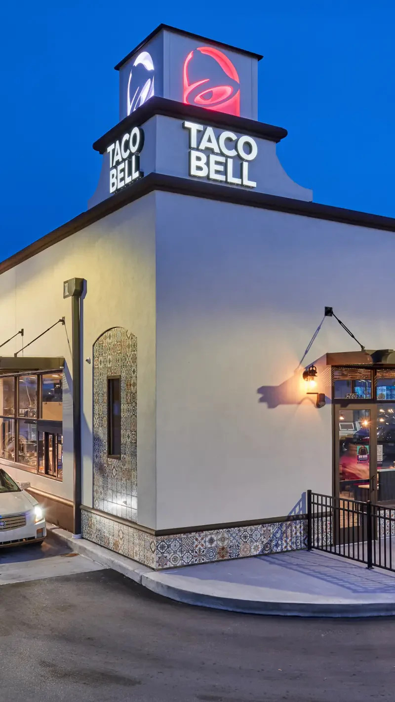 A Taco Bell restaurant is shown in the image, featuring its iconic logo illuminated against a twilight sky. The building showcases modern architecture with decorative tiles.