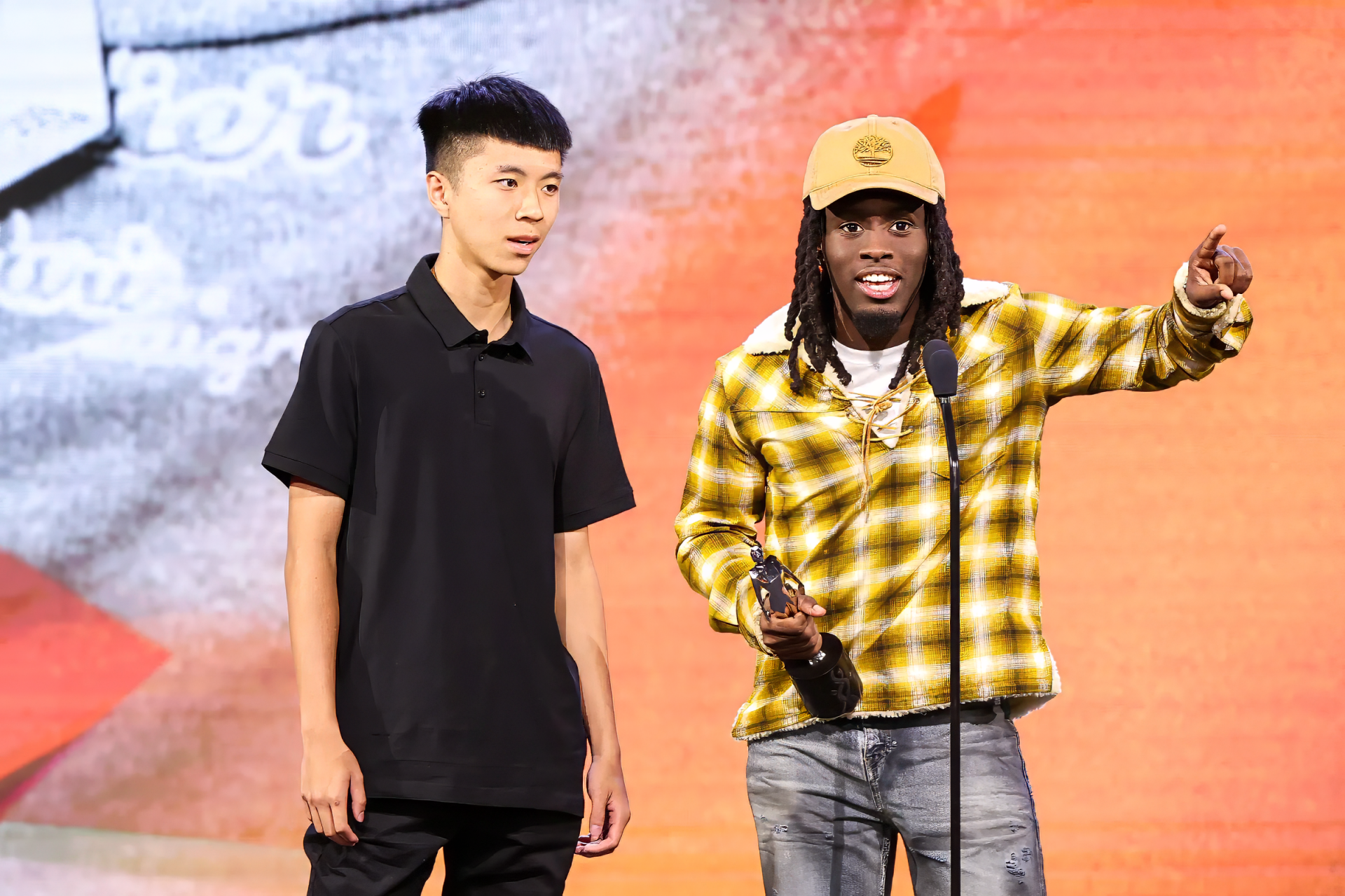 Kai Cenat, a well-known streamer, gestures while accepting an award on stage, accompanied by another individual. The backdrop features vibrant colors and patterns.