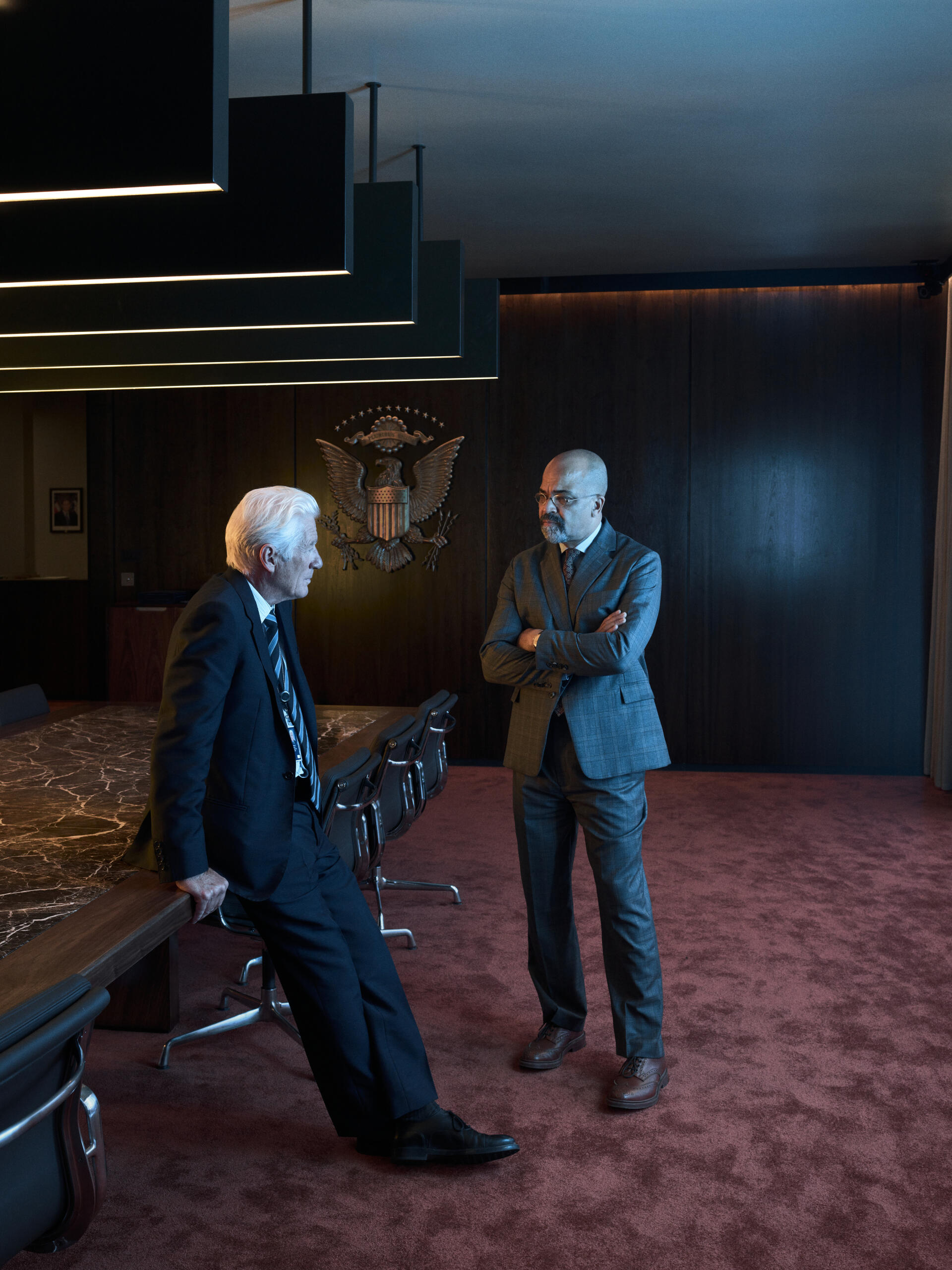 Richard Gere and Jeffrey Wright engage in conversation in a sleek, modern boardroom setting from the TV show The Agency. The ambiance is professional and tense.