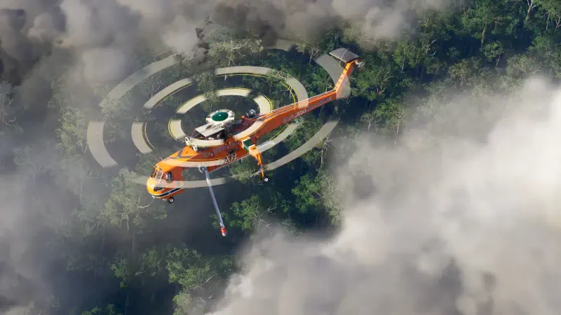 An orange helicopter hovers above a forest, surrounded by clouds, showcasing the detailed environments of Microsoft Flight Simulator 2024.