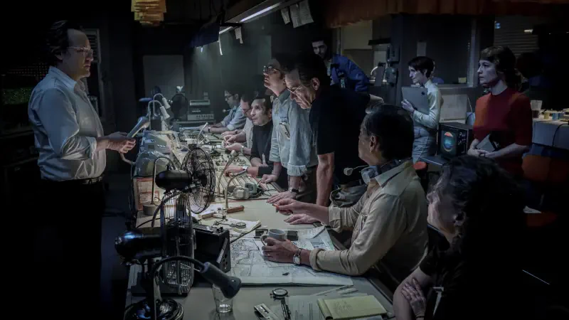 A group of people gathers around a cluttered table in a dimly lit room, attentively listening to a speaker, conveying a sense of intrigue and collaboration. Movie release date: September 5.